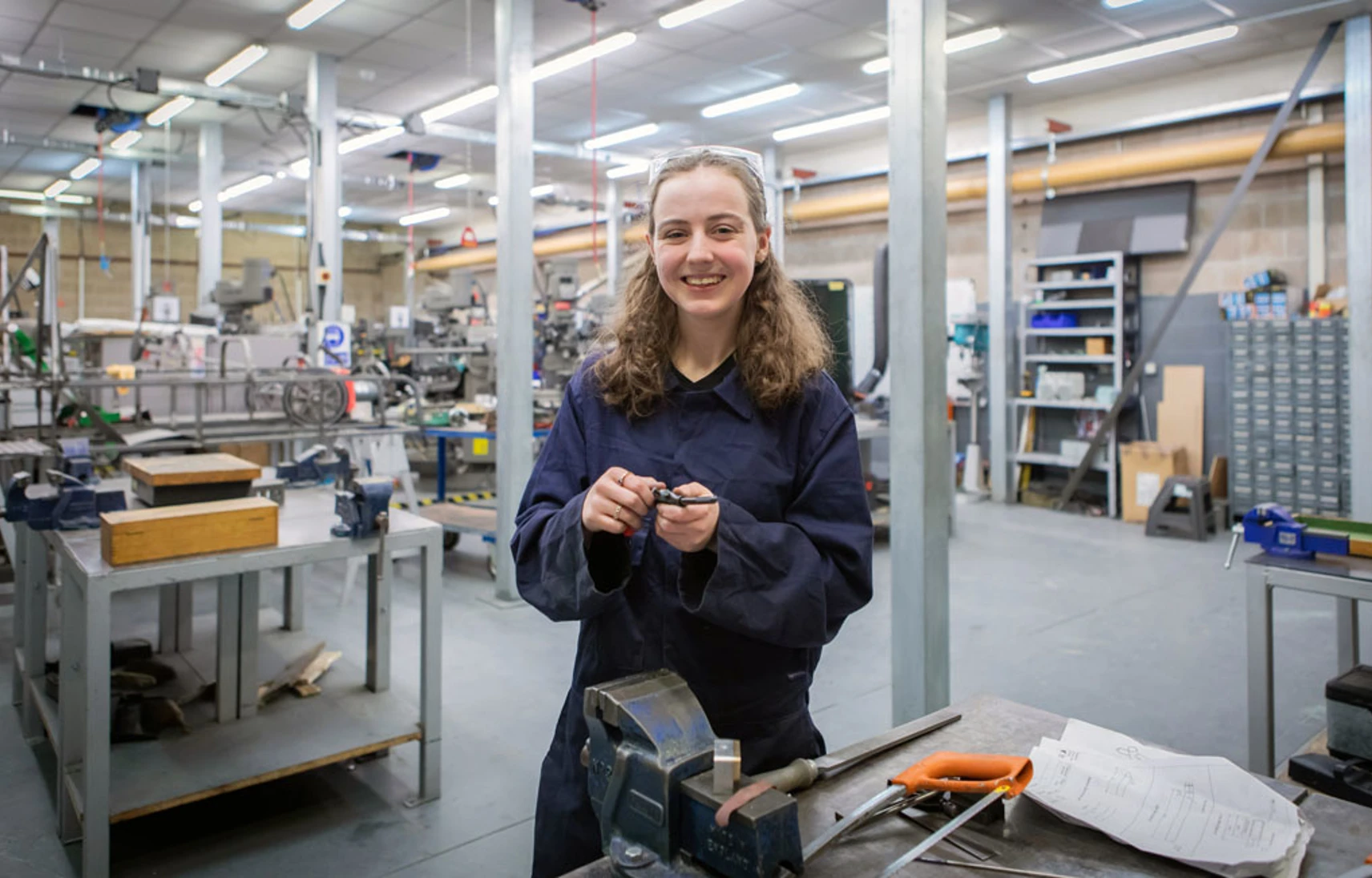 A female mechanical engineering student in navy-blue overalls smiling while working on a small metal component in a busy college workshop. A female mechanical engineering student in navy-blue overalls smiling while working on a small metal component in a busy college workshop.